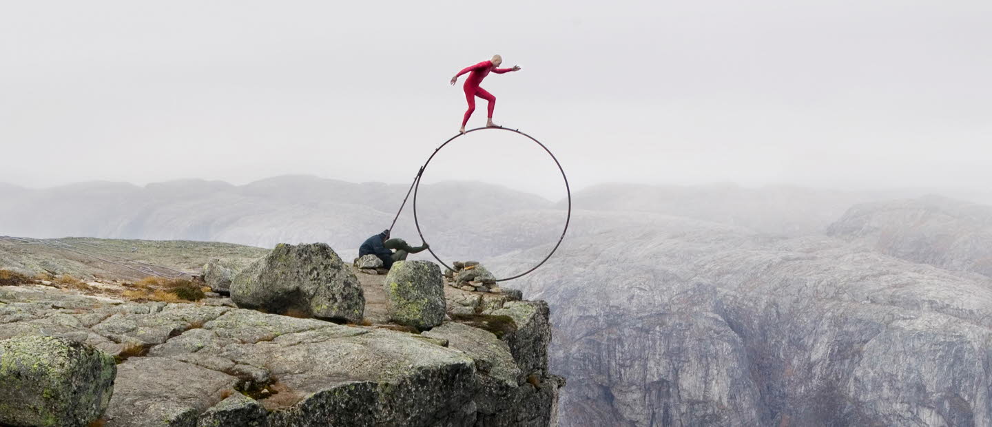 En man klädd i rött står på en stor, smal rund ring precis vid kanten av ett berg.