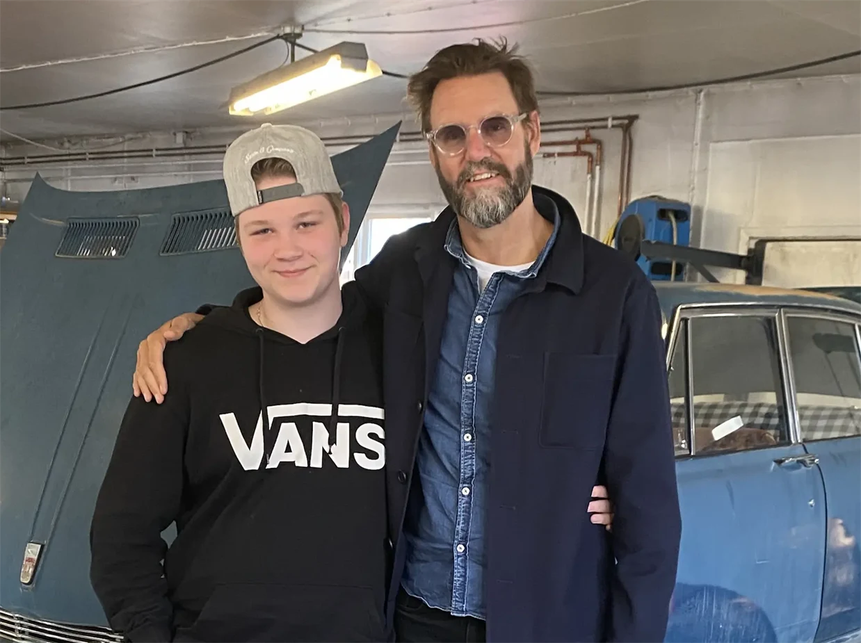 En ung kille står tillsammans med en författare framför en bil med öppen motorhuv.
