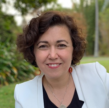 A woman in a white suit jacket is looking into the camera. She has brown hair and smiling.
