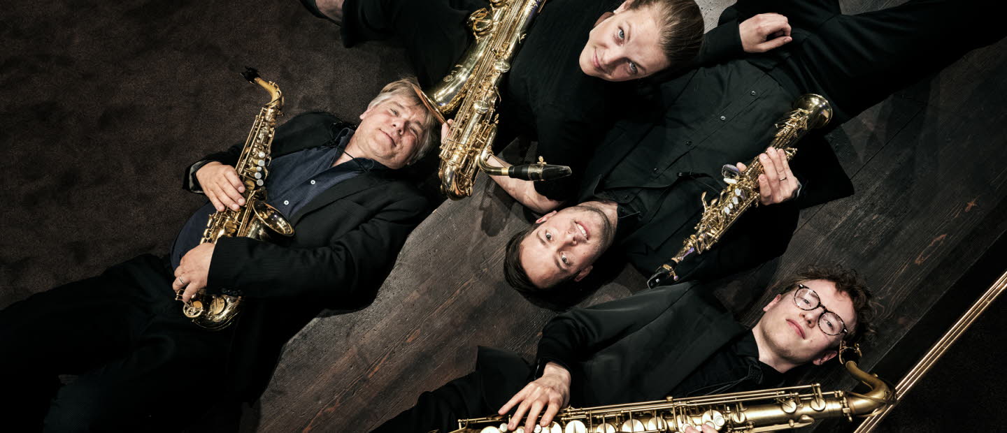 Fyra musiker klädda i svart ligger på golvet, håller blåsinstrument i famnen. 