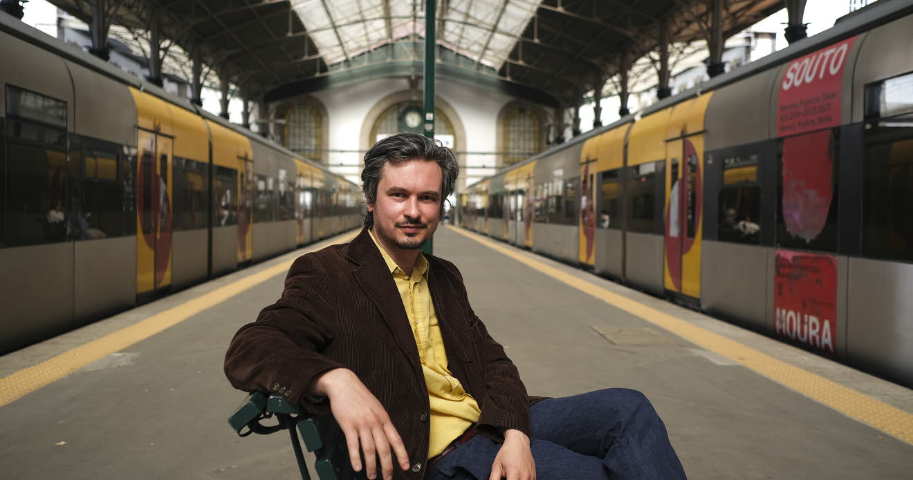 Bild på Maxim på en tågperrong med två tåg bredvid. Han sitter på en stol med gul skjorta och mörk kavaj. 