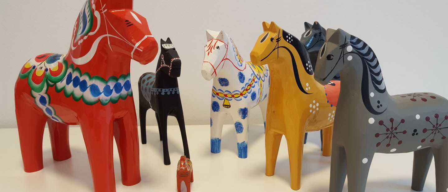 Flera dalahästar i olika storlekar och färger stående på ett vitt underlag.