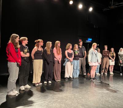 En rad ungdomar står på en scen, en ung kvinna står lite längre fram vid en mikrofon.