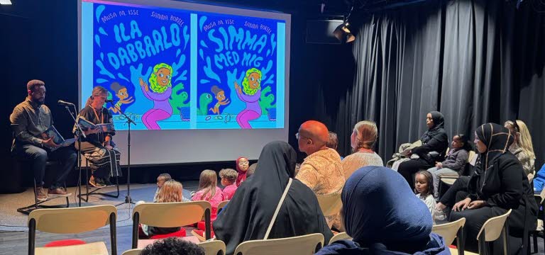 Två personer sitter på en scen och läser framför en publik med barn och vuxna.