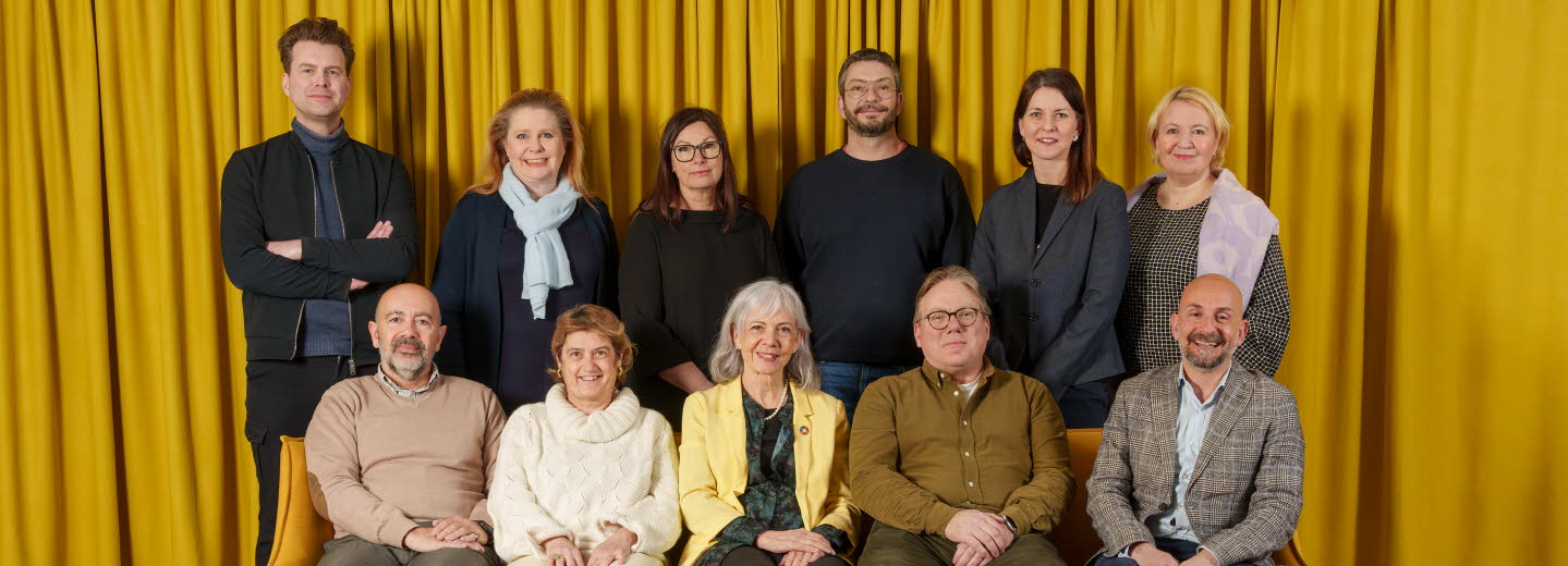 Gruppbild på jurymedlemmar, fem personer sitter i en gul soffa, bakom soffan står sex personer, i bakgrunden hänger en stor gul gardin. 