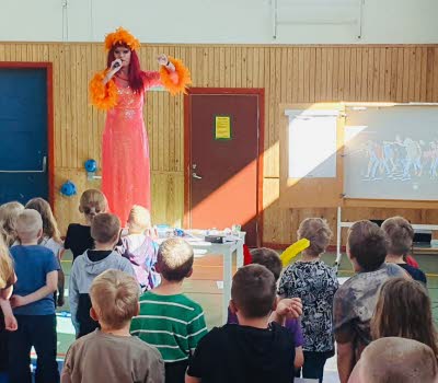 Dragqueen artisten Inga Tvivel står på en scen och undervisar en skolklass. 
