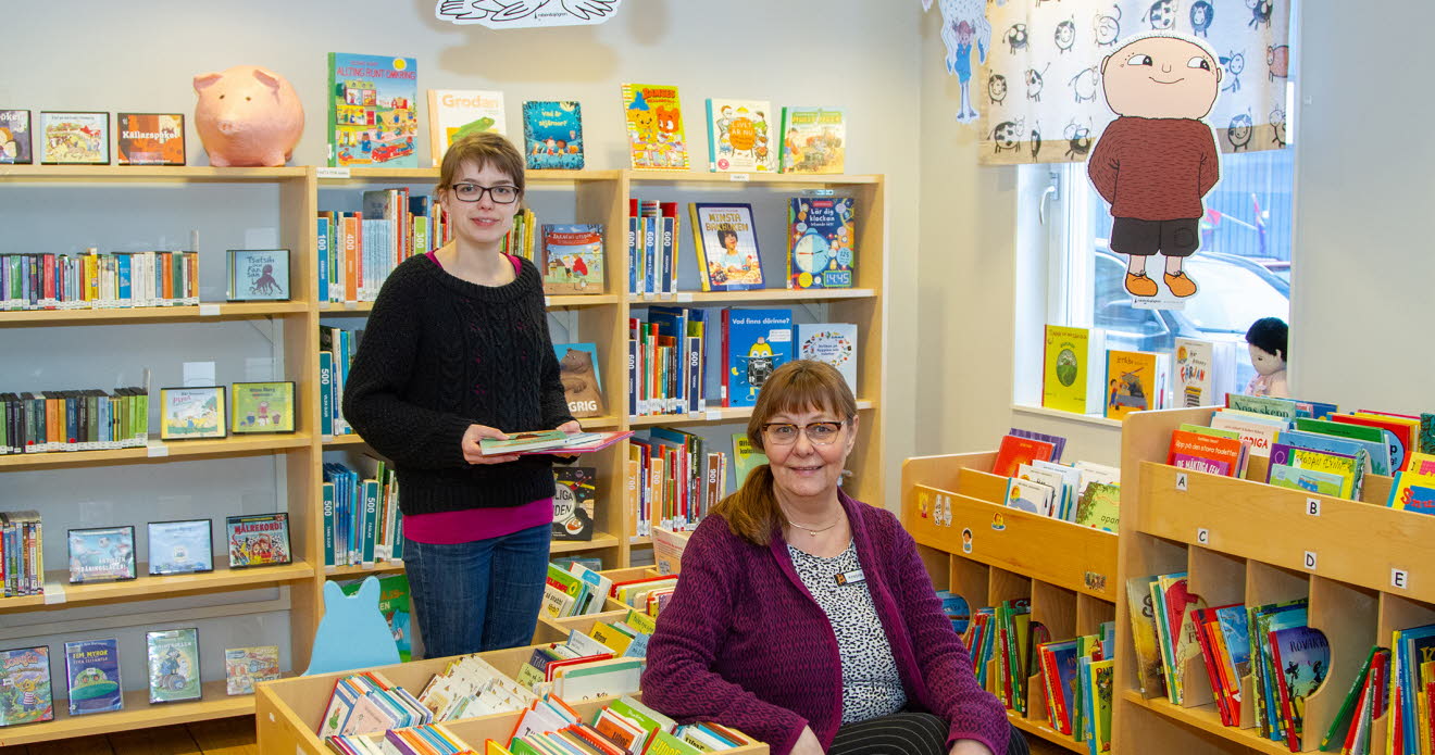 Två kvinnor, en sittande och en stående, står i bibliotekets barnboksområde. En bild av Alfons Åberg hänger vid fönstret.