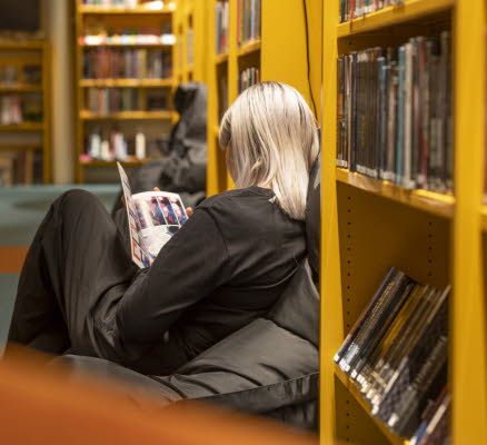Tonårstjej sitter i fåtölj på bibliotek och läser en bok.