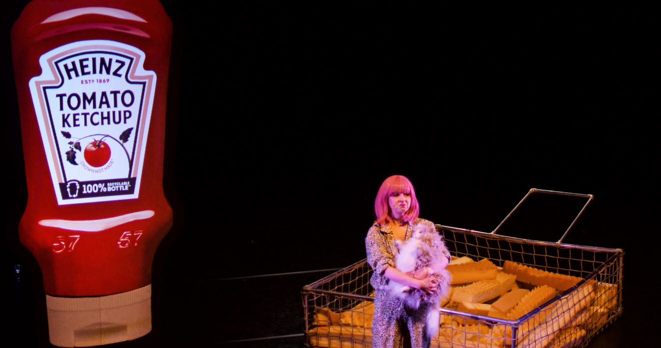 En tjej i rosa hår står på en mörk scen, i bakgrunden syns en förstorad ketchupflaska och förstorade pommes frites..
