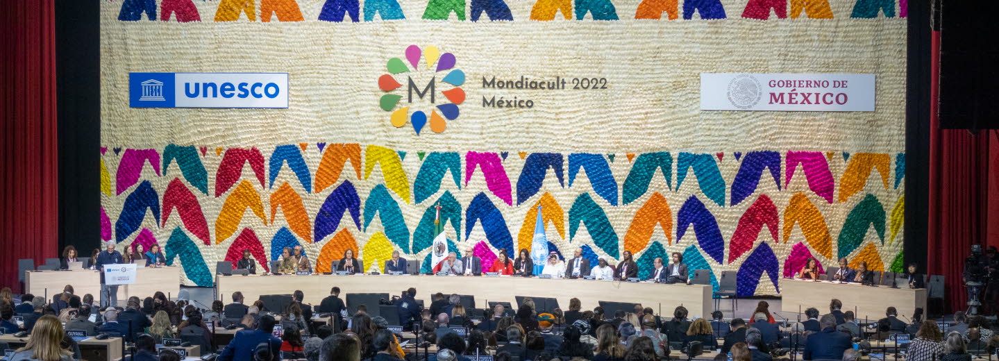 Publik som sitter framför scenen. Scenen har en vägg fylld med loggor och färgglada illustrationer. 