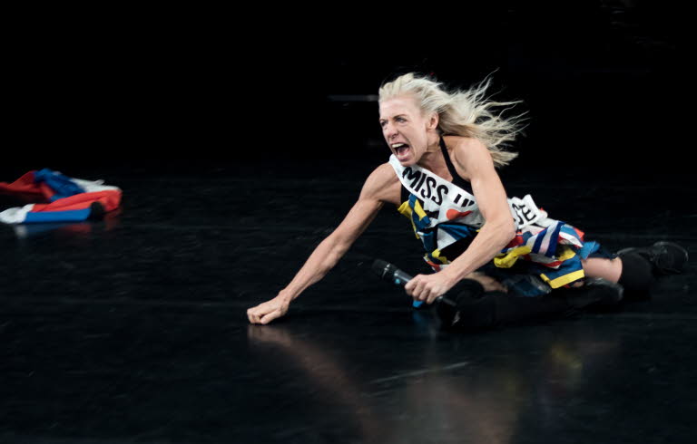 En kvinna med mikrofon halvliggandes på ett scengolv.