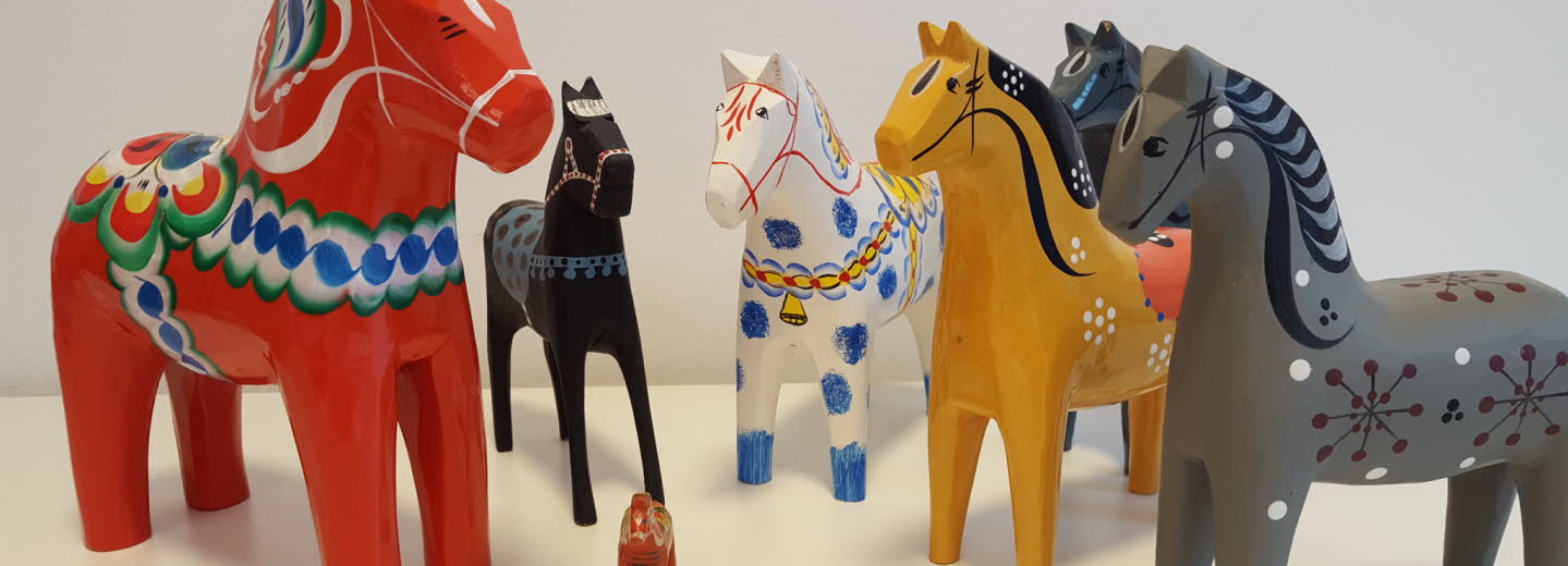 Flera dalahästar i olika storlekar och färger stående på ett vitt underlag.