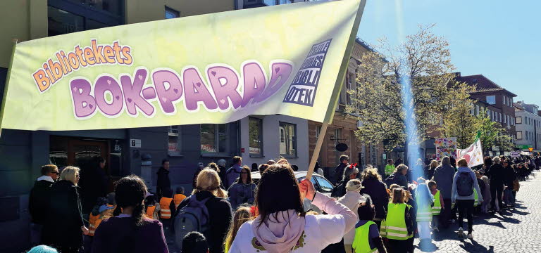 Barn och vuxna som tågar längs en gata med banderoller med texten Bibliotekets bokparad.