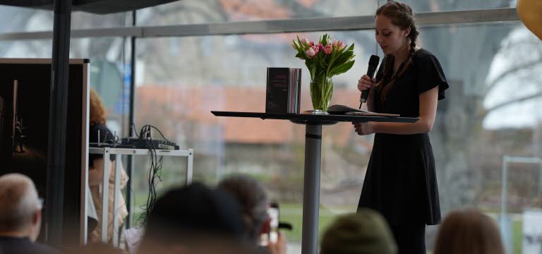 En ung kvinna står med en mikrofon på en scen och läser ur en bok.