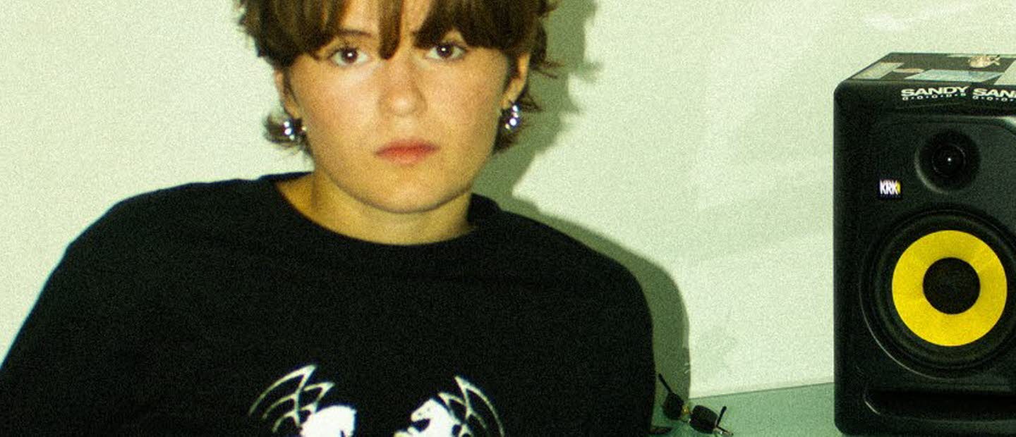 Kvinna i 20-årsåldern sittandes vid ett bord framför en laptop. På bordet en högtalare och hörlurar. 