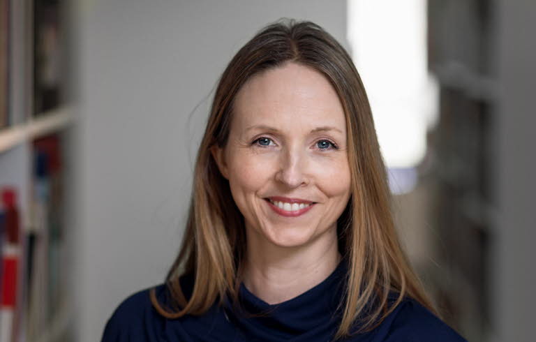 Medelålders kvinna med långt cendréfärgat hår och blå tröja omgiven av bokhyllor.