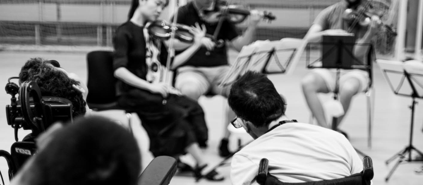 Personer i rullstol lyssnar på ett liveframförande av klassisk musik. Bilden är i svart-vitt och tillsynes taget i en gympasal.