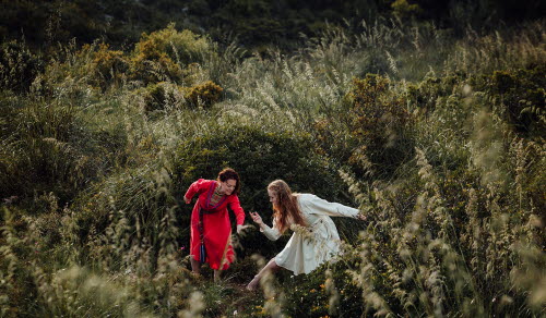 Två kvinnor, den ena i röd dräkt och den andra i vit, danser och jojkar omgivna av högt gräs med vita plymer.