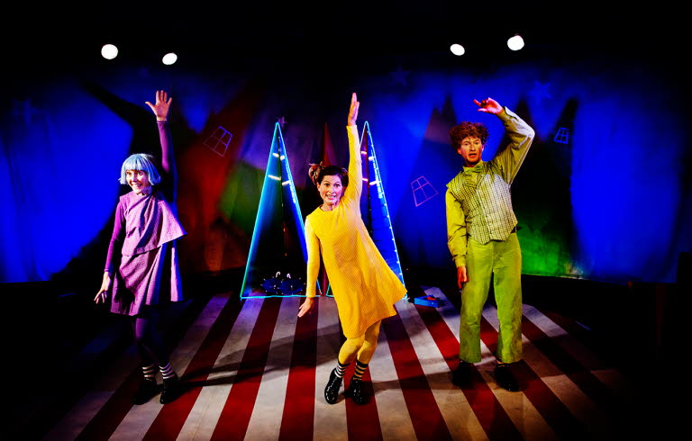 Tre vuxna som mimar och dansar. Färgstark scenografi med bredrandigt golv och blåbelysta väggar.
