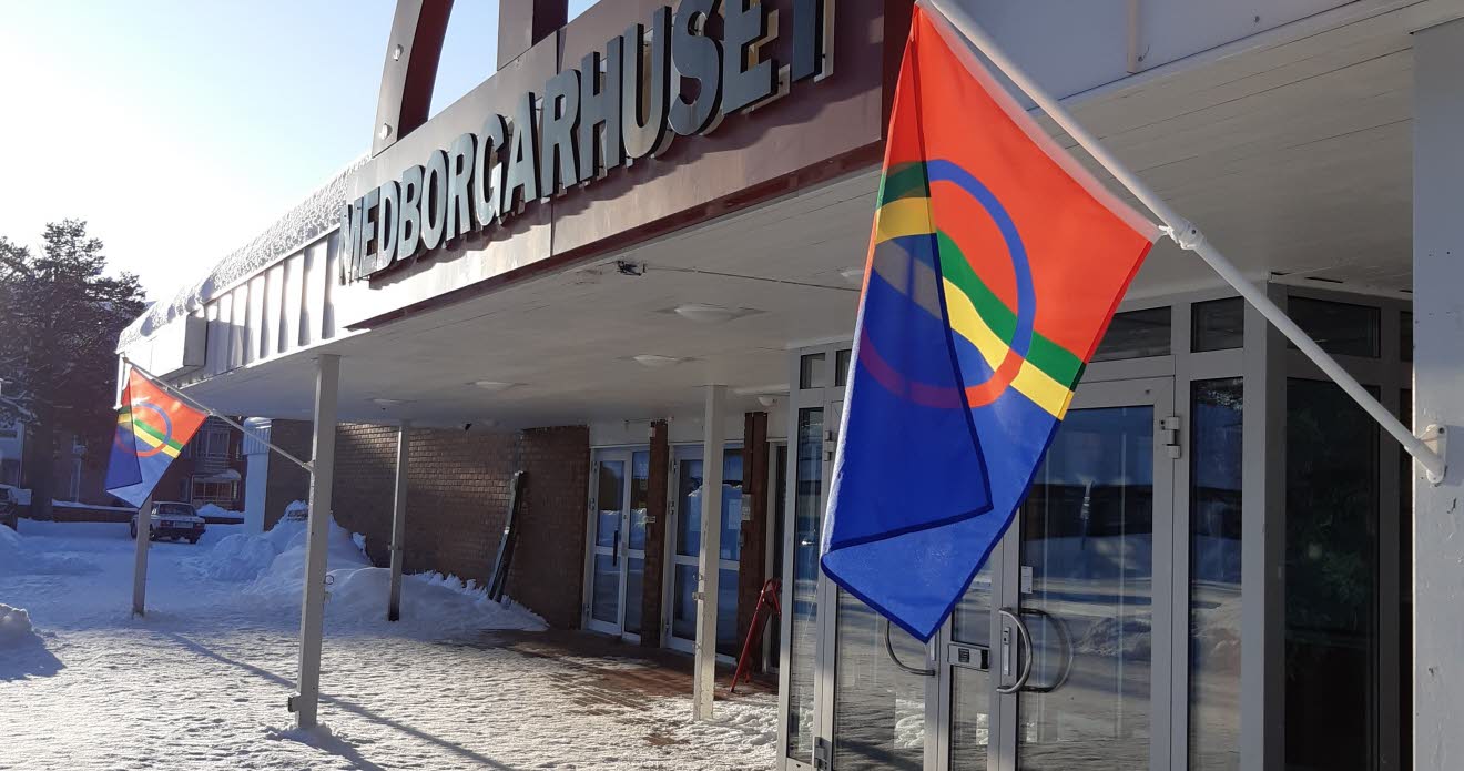 Exteriör: En solig vinterdag utanför Medborgarhuset. Två samiska flaggor hänger vid ingången. Det är snö på marken. 