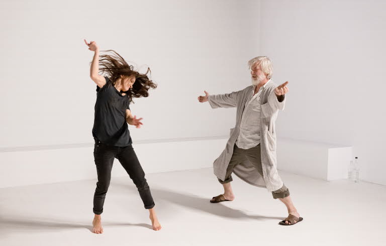 A young woman and an old man are doing a modern dance in a white room.