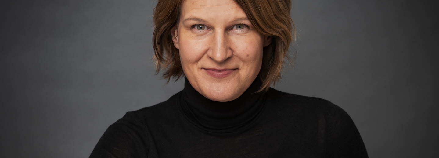 Porträttbild av medelålderskvinna som ler och tittar in i kameran. Mörkt axellångt hår, svart polo, mörkgrå bakgrund. 