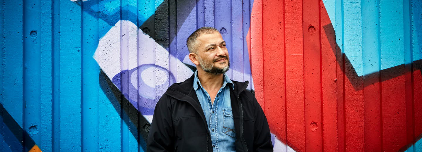 Bagir Kwiek står framför en vägg med graffiti och ler. Han tittar snett ut ur bild. 