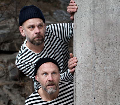 Anders Sparring and Per Gustavsson, dressed in black and white sweaters/prison style, looking out behind a wall.