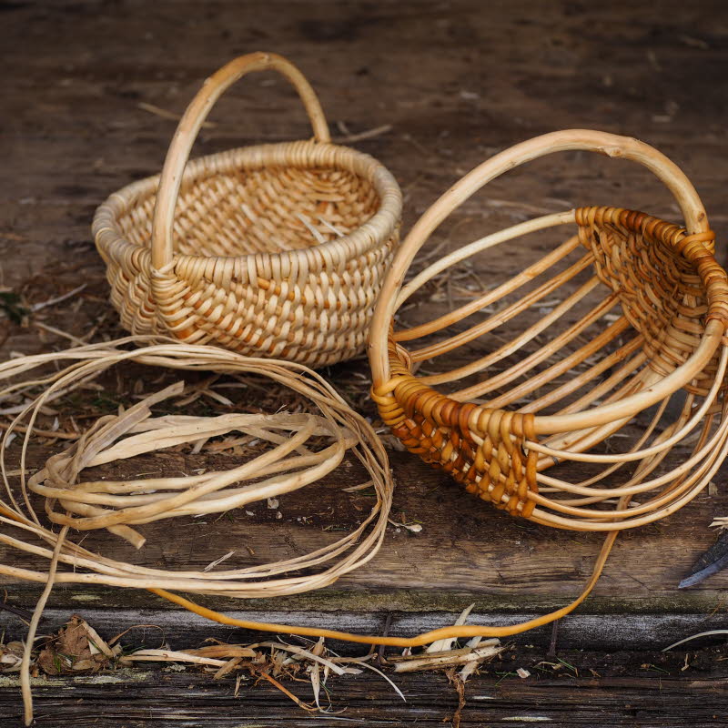 Två flätade korgar i naturmaterial, omgivna av rötter och en kniv på ett träbord.