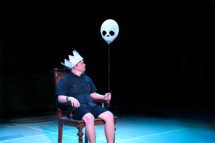 En man sitter på en scen. På huvudet har han en kungakrona i papp, i handen håller han en ballong med spökansikte.