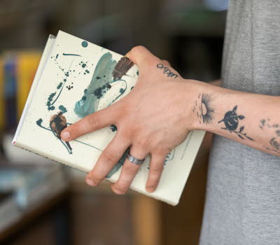 Närbild på tatuerad arm, handen håller i en bok.
