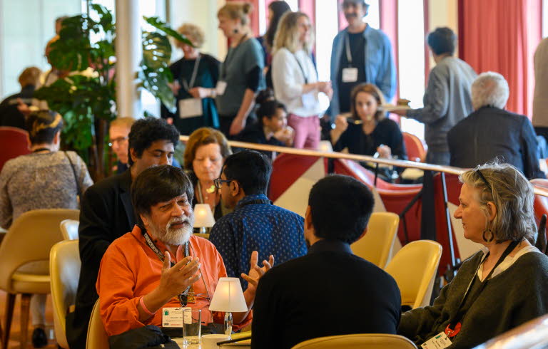 Fullsatt rum med människor som pratar i sittgrupper runt olika bort.