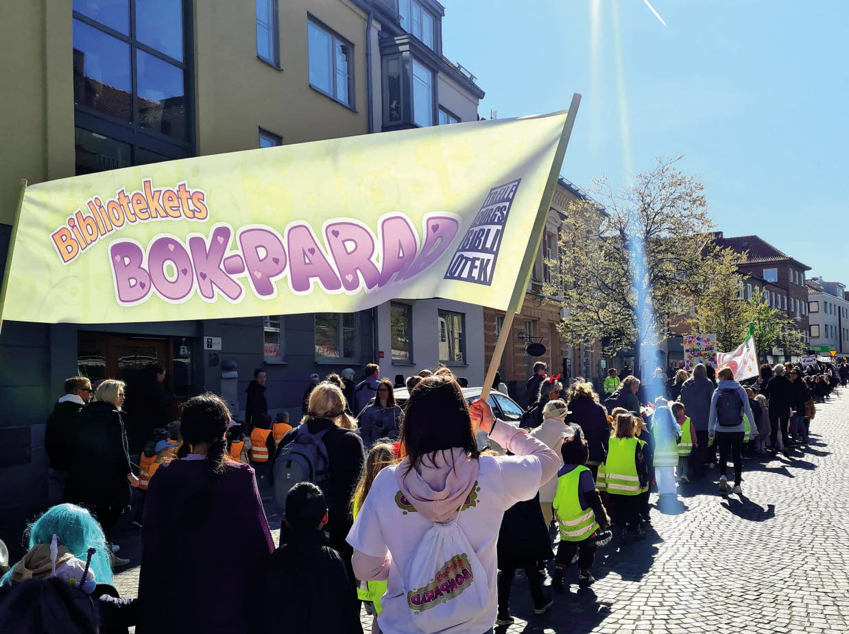 Barn och vuxna som tågar längs en gata med banderoller med texten Bibliotekets bokparad.