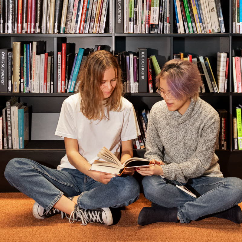 Närbild på bokhyllor fyllda med böcker. Två barn sitter på golvet.