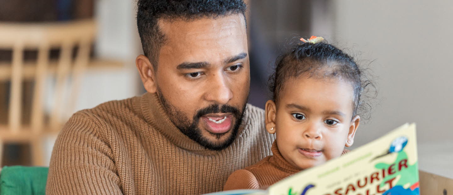 En liten flicka sitter i en mans knä som pekar och läser ur en bok om dinosaurier. Flickan tittar koncentrerat i boken. 