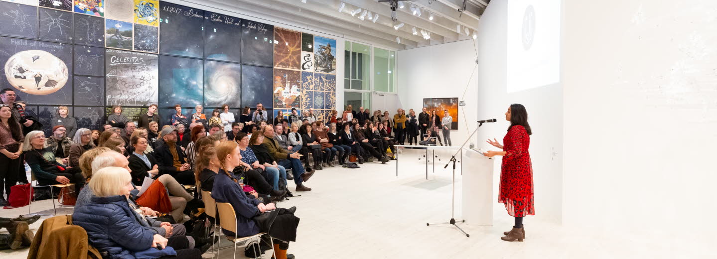 En kvinna som står på vid en talarpulpet och talar till en stor publik. 