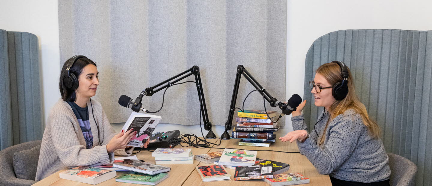 Två biblioteksmedarbetare sitter i en poddstudio med massa böcker som de diskuterar. 