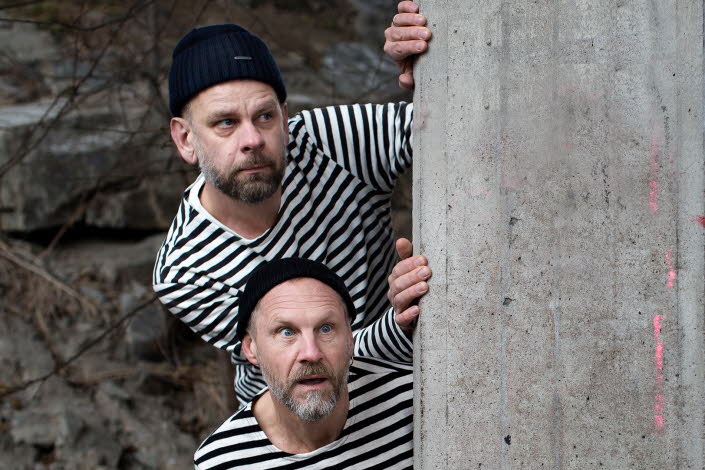 Anders Sparring and Per Gustavsson, dressed in black and white sweaters/prison style, looking out behind a wall.