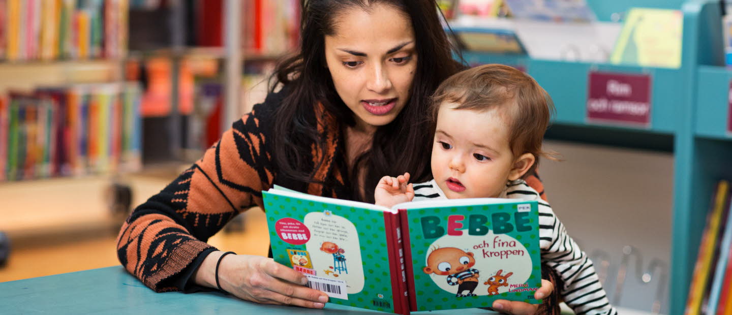 Kvinna och barn sitter vid ett litet bord på ett bibliotek och titta i en pekbok.