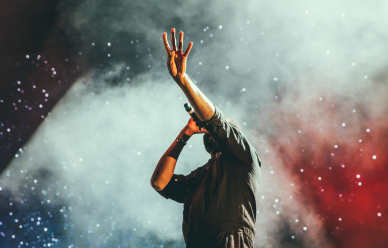 Man som sjunger i mikrofon med vänstra armen upp i luften. I bakgrunden syns rök i vitt och rött samt ljus som påminner om stjärnor.
