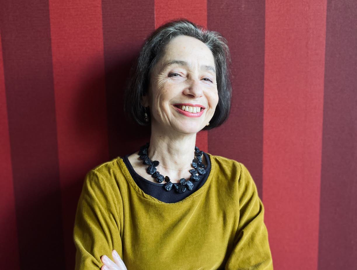 Foto på Elena Balzamo som ler mot kameran. Elena har en mörk page och är klädd i en senapsgul klänning med ett stort svart halsband. Bakgrunden är randig i röda toner. 