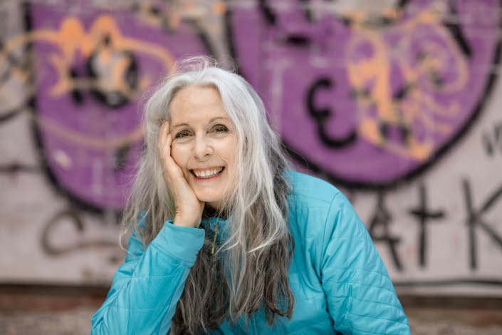 Portrait of Laurie Halse Anderson sitting in front of a graffiti painting. She's smiling and looking towards the camera. 