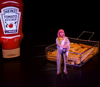 En tjej i rosa hår står på en mörk scen, i bakgrunden syns en förstorad ketchupflaska och förstorade pommes frites..
