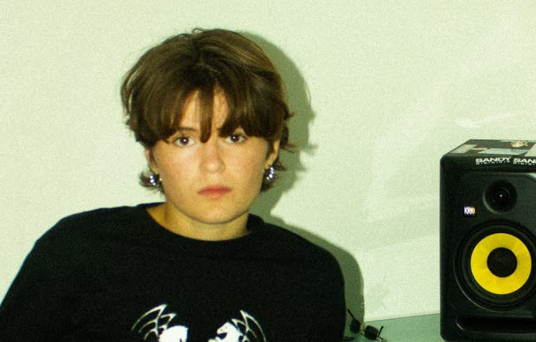 Kvinna i 20-årsåldern sittandes vid ett bord framför en laptop. På bordet en högtalare och hörlurar. 