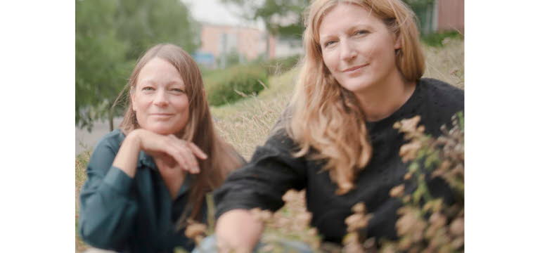 Foto på Jenny och Lotta som sitter ner i gräset, i bakgrunden skymtas träd och en tegelbyggnad.