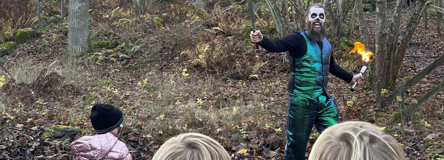 Barn som tittar på en artist i grönblank kostym med en fackla i handen ute i skogen. 