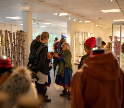 Två besökare samtalar med en samiskt klädd kvinna. De står i en vit lokal där kläder hänger på skyltdockor. Till vänster om dem står nedhuggna björkstammar uppradade. 