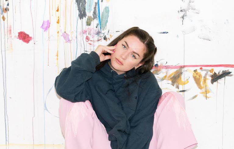 Artist Moa Romanova is sitting against a wall in pink pants and a black hoodie.
