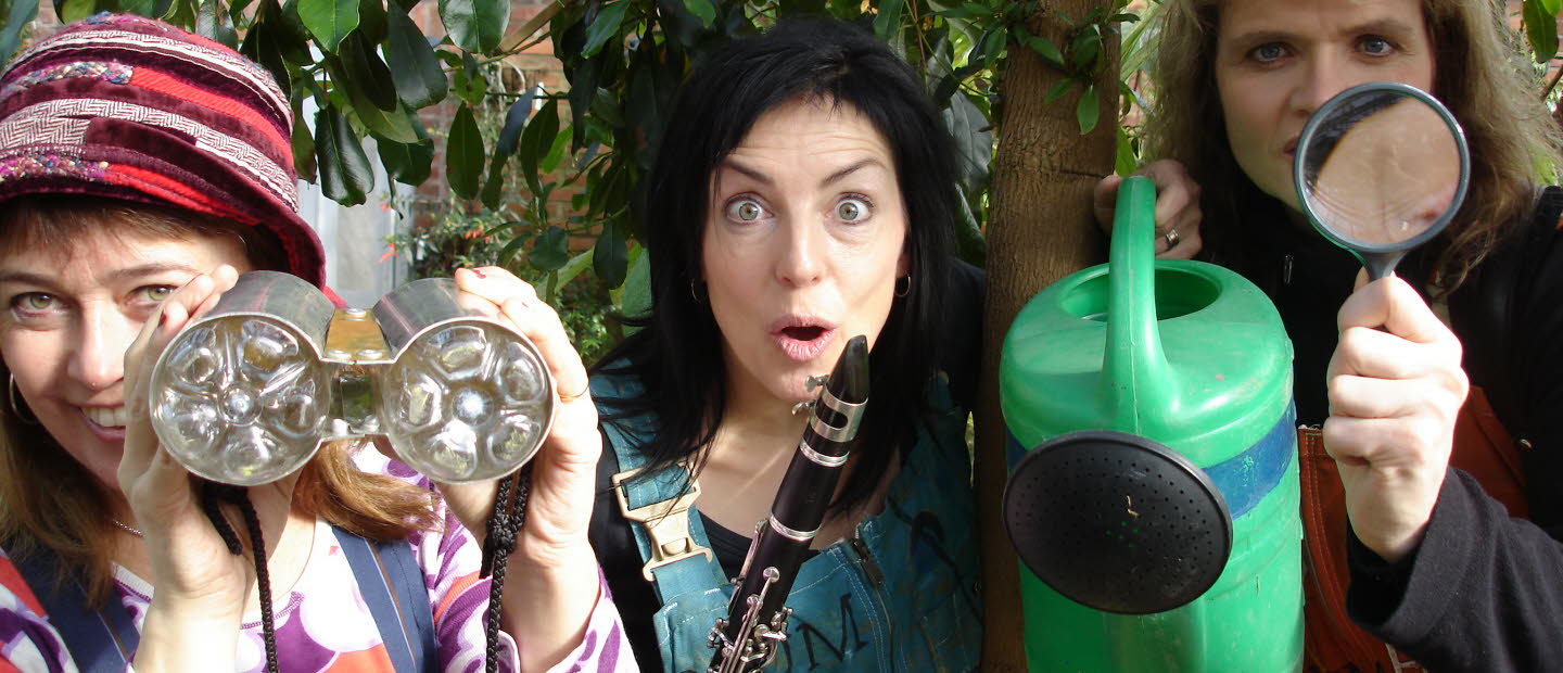 Tre personer står bland träd och tittar nyfiket in i kameran. De håller i en vattenkanna, en flöjt och en kikare. 