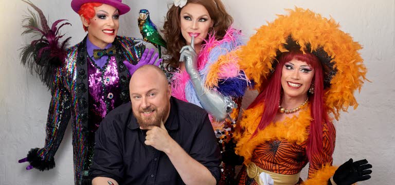 En person sitter vid ett skrivbord och skriver, bakom syns tre dragqueens.